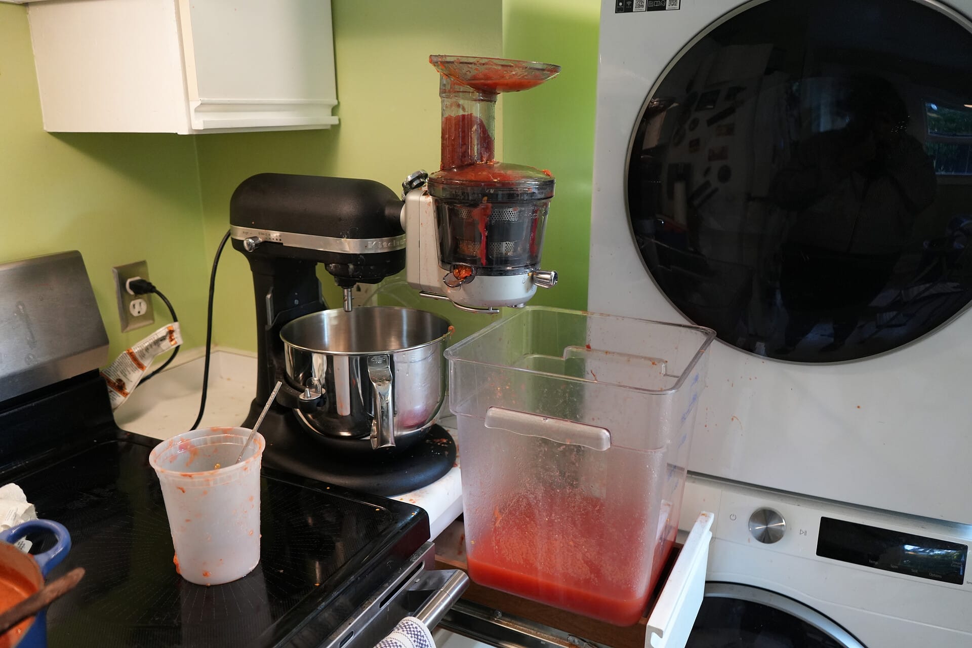 Slow juicer attachment for a Kitchenaid stand mixer flowing into a 18-quart bin with some tomato juice at the bottom.